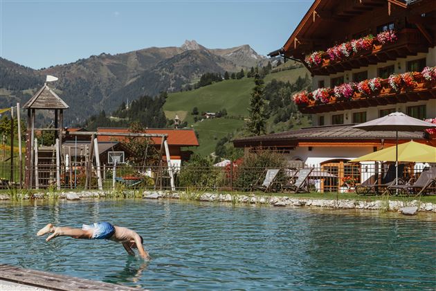 A man jumps into a clear, blue pool in the mountains. In the background, there is a building with flower balconies and a green landscape.