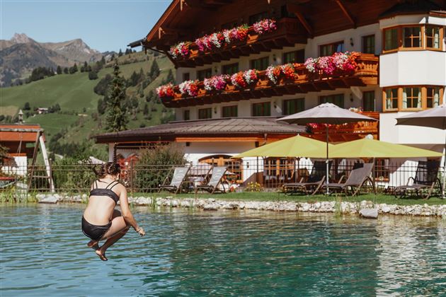 Eine Person springt in einen Pool vor einem hübschen Hotel mit blühenden Balkonpflanzen. Im Hintergrund sind grüne Berge zu sehen.