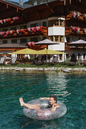 Ein Mädchen entspannt auf einem aufblasbaren Reifen im klaren Wasser. Im Hintergrund ist ein schönes Hotel mit blühenden Pflanzen und Sonnenschirmen zu sehen.