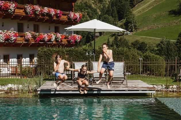 Three children are getting ready to jump into a clear pool. In the background, there is a beautiful house and green hills visible.