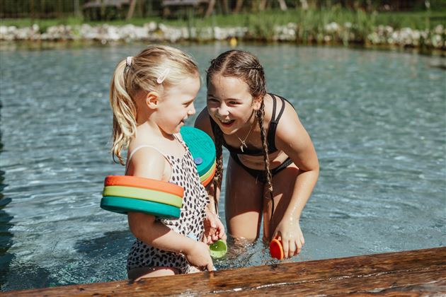 Zwei fröhliche Mädchen spielen im Wasser. Sie tragen Schwimmhilfen und lachen zusammen.