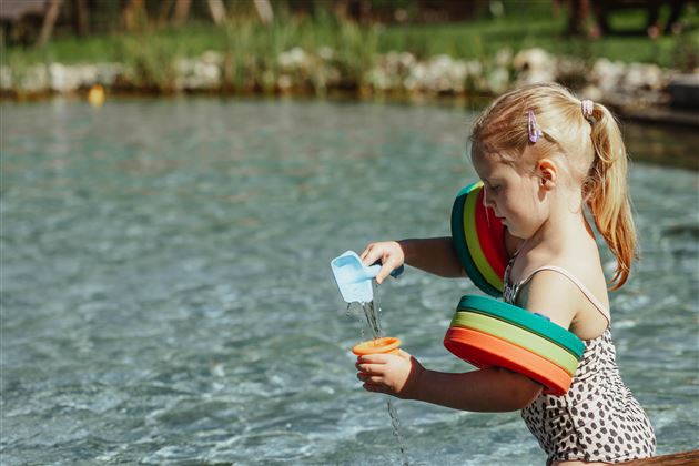 Ein Mädchen spielt im Wasser und gießt mit einem kleinen Behälter. Sie trägt bunte Schwimmflügel und hat blondes Haar.