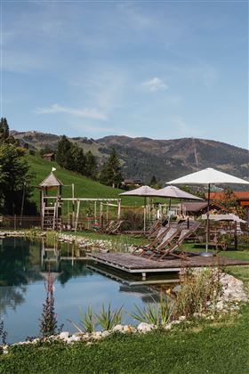 Ein idyllischer Poolbereich mit Liegen, umgeben von grünen Wiesen und Bergen. Im Hintergrund sind Sonnenschirme und Spielgeräte zu sehen.