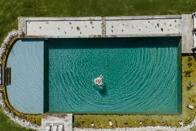 A beautiful pool with clear water and a person swimming in it. The surroundings are green and well-kept, with stones and grass.