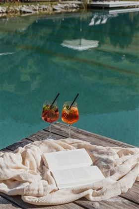 Two cocktails are on a wooden table next to an open book. In the background, the clear turquoise water is reflected.