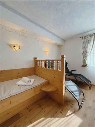 A cozy room with a wooden bed and a reclining chair.  
Soft lighting and bright curtains create an inviting atmosphere.