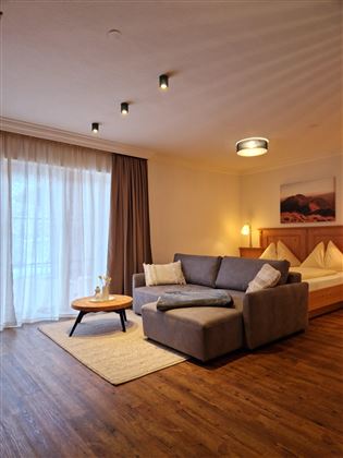 Ein modernes Zimmer mit einem gemütlichen Sofa, einem kleinen Tisch und einem großen Bett im Hintergrund. Die Fenster sind lichtdurchflutet und verleihen dem Raum eine einladende Atmosphäre.