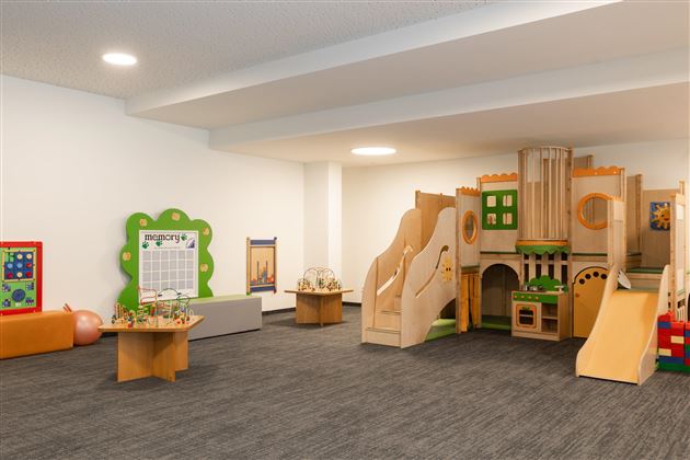 Ein einladender Spielraum mit Holzspielgeräten, Rutschen und einem künstlerischen Bereich. Der Raum ist hell und freundlich gestaltet, ideal für Kinder zum Spielen und Entdecken.