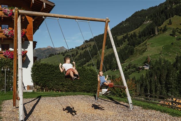Zwei Kinder schaukeln fröhlich auf einer Spielplatzschaukel in einer malerischen Landschaft. Im Hintergrund sind grüne Hügel und ein bewaldeter Hang zu sehen.