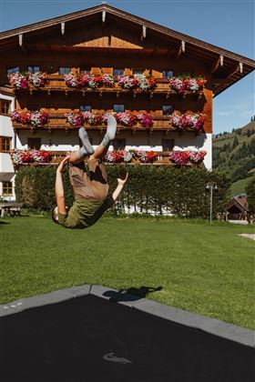 Ein Junge springt auf einem Trampolin und macht einen Rückwärtssalto. Im Hintergrund steht ein Holzhaus mit bunten Blumen an den Fenstern.