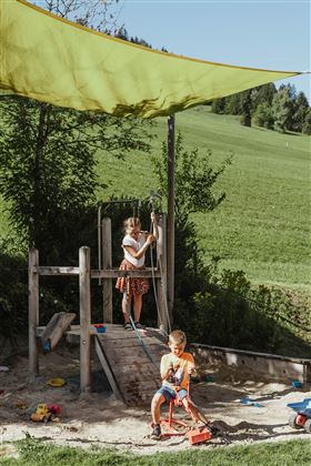 Two children are playing on a playground under a sunshade. In the background, green meadows can be seen.