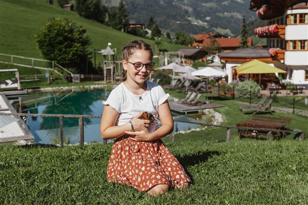 Ein Mädchen sitzt auf dem Gras und hält ein kleines Tier in den Händen. Im Hintergrund ist ein schöner Pool und eine grüne Landschaft zu sehen.