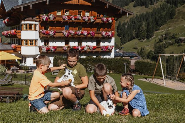 Vier Kinder sitzen auf der Wiese und spielen mit Zwergkaninchen. Im Hintergrund ist ein schön dekoriertes Gebäude mit Blumen vor einer grünen Berglandschaft zu sehen.