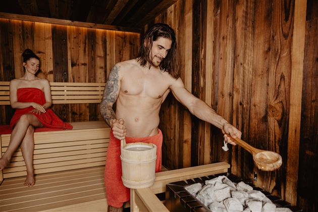 A man in a sauna pours water on hot stones while a woman relaxes in a red towel. The walls are made of wood, and the atmosphere is cozy and relaxing.