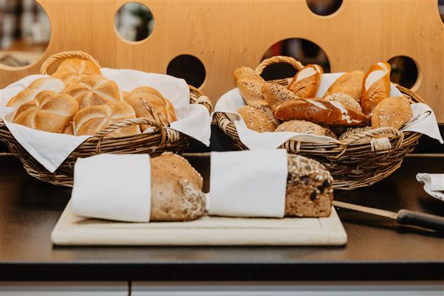 A basket with fresh bread and various rolls is on a table. Next to it, a sliced bread is lying on a board.