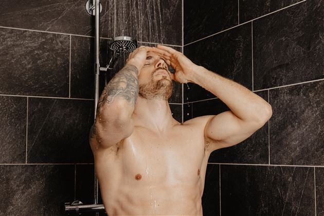 A man is standing under the shower, washing his hair. The walls are made of dark tiles and water is flowing over his body.