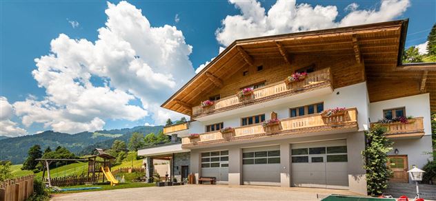 Ein charmantes Landhaus mit einem Holz-Dach und Balkonblumen. Im Hintergrund sind grüne Hügel und ein blauer Himmel mit einigen Wolken zu sehen.