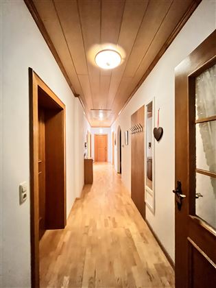 Ein schmaler Flur mit Holzfußboden und weißen Wänden. An der linken Seite sind mehrere Türen und ein Wandhaken zu sehen.