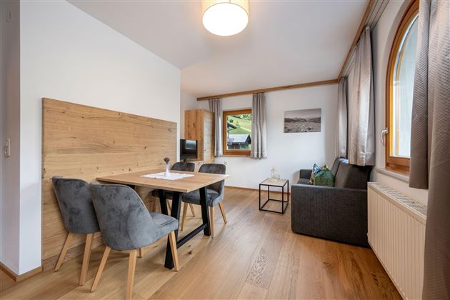 A modern living room with a dining table, chairs, and a cozy couch. Large windows let in plenty of light and offer a view of the surroundings.