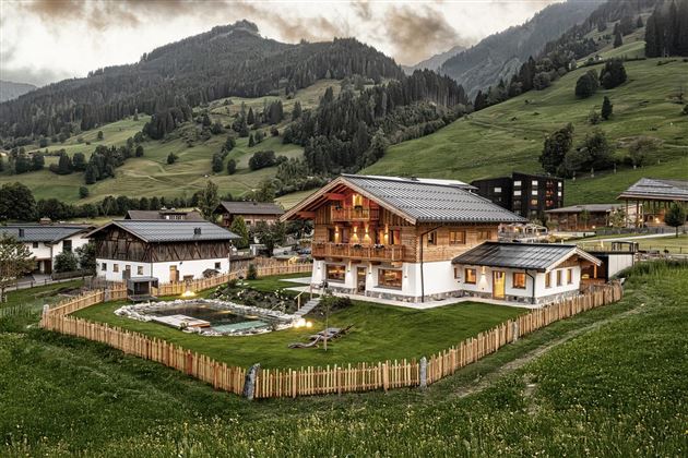 Ein schönes, alpenländisches Haus umgeben von grünen Wiesen und Bergen. Der Garten ist gut gepflegt und von einem Zaun umgeben.