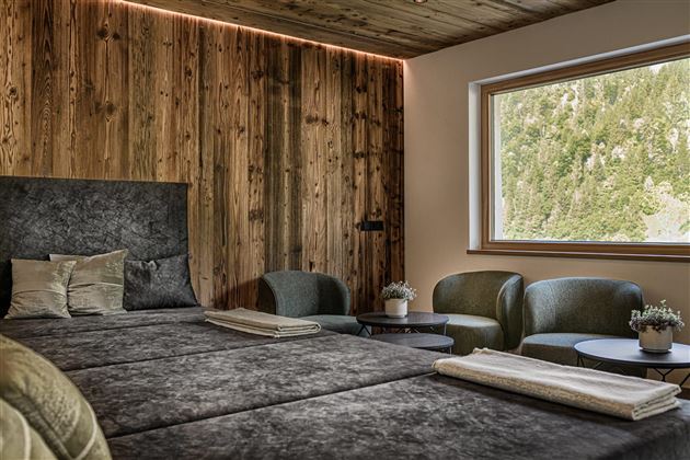 A modern bedroom with a large bed and cozy armchairs. The walls are made of wood and there is a window overlooking nature.