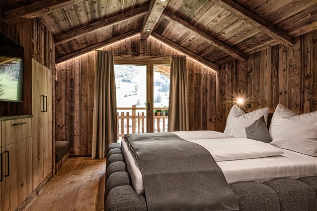 A cozy bedroom with a wooden ceiling and warm light. The room has a large window with a view of the mountains.