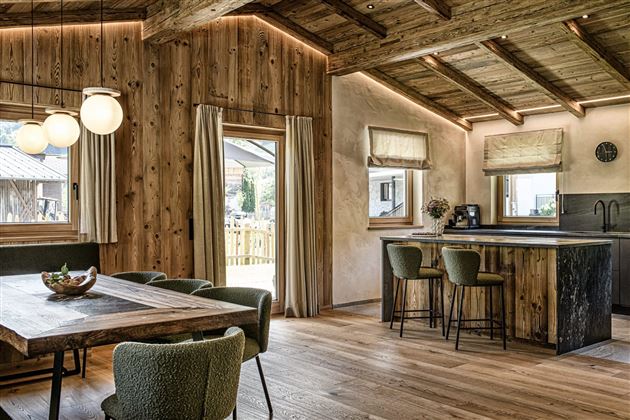 A modern, cozy living room with wood paneling and a large dining table. The room has large windows that let in plenty of daylight.