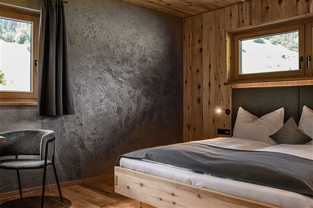 A modern bedroom with wooden details and large windows. There is a comfortable bed and a table with a chair.
