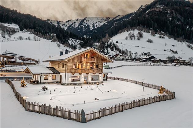 A cozy wooden house in a snowy landscape. The surroundings are surrounded by mountains, and the garden is enclosed by a wooden fence.