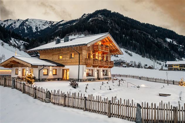 A charming wooden house in winter with a snow-covered garden. In the background, the mountains and a cloudy sky can be seen.