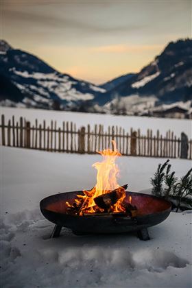 A cozy fire in a bowl stove stands on the snow. In the background, snowy mountains and a wooden fence can be seen.