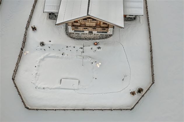 A cozy chalet in the snow, surrounded by a wooden fence. The garden shows traces in the white snow and some lights.