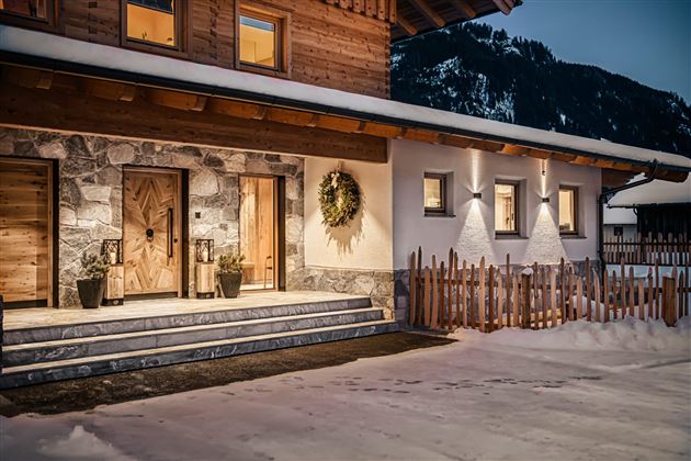 A cozy wooden house in the snow with an inviting front door. The facade is modern and stylish, surrounded by mountains and a tranquil winter landscape.