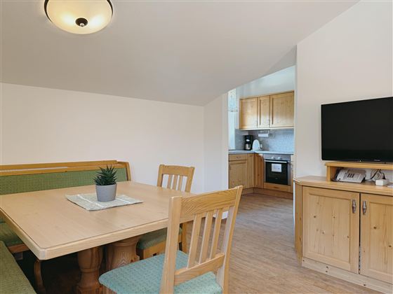 A cozy dining area with a wooden table and chairs. In the background, you can see a modern kitchenette and a television.