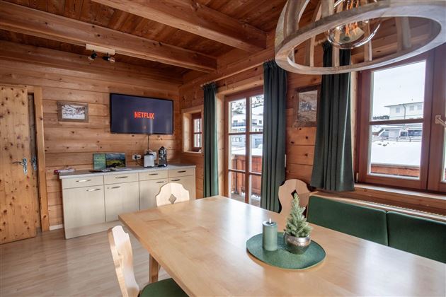 Ein gemütlicher Holzraum mit einem Esstisch und Stühlen. An der Wand hängt ein Fernseher, und große Fenster lassen viel Licht herein.