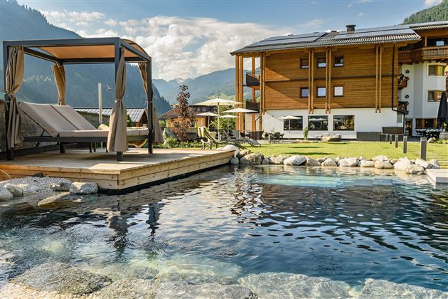 A peaceful garden with a glittering pool and a cozy lounger. In the background stands a modern wooden house in front of a picturesque mountain landscape.