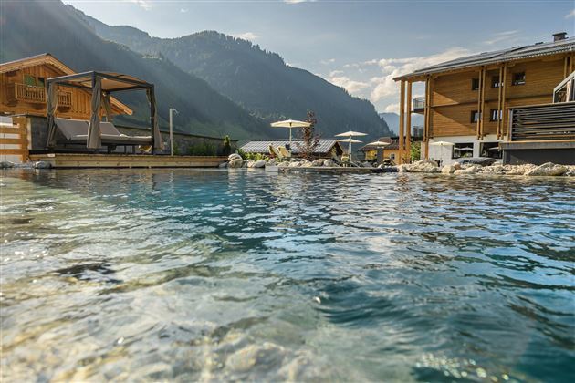 A pool with clear water, surrounded by pretty wooden cabins in a mountainous landscape. In the background, the gentle mountains and a blue sky are visible.