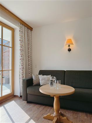A cozy living room with a green sofa and a round table. A lamp hangs on the wall, and a window lets in plenty of daylight.