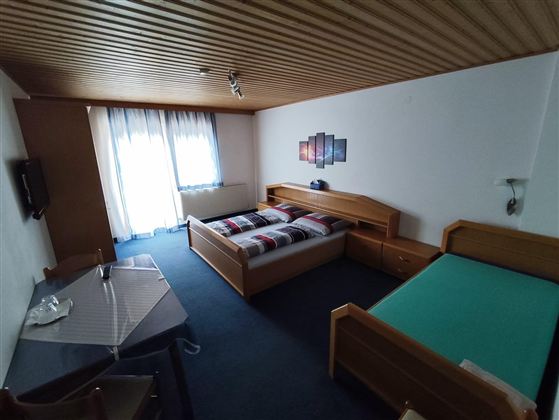 Ein einfaches Zimmer mit zwei Betten und einem Tisch. Das Zimmer hat einen Fernseher und große Fenster, die viel Licht hereinlassen.