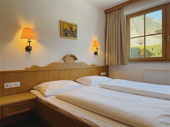 A cozy room with two separate beds and warm light. The window offers a view of greenery.