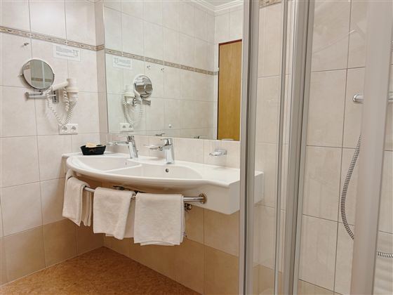 A modern bathroom with a double sink unit and towels. The walls are brightly tiled, and there is a shower in the background.