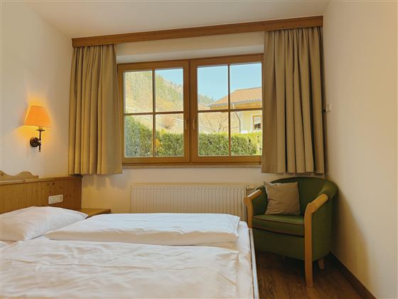 A cozy room with two beds and an armchair. Large windows offer a view of the green landscape.