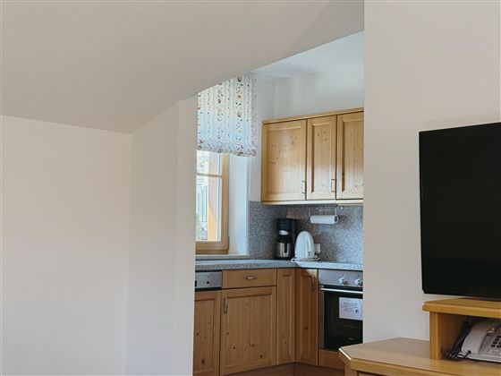 A bright kitchen with wooden cabinets and modern appliances. In the background, a window and a television are visible.