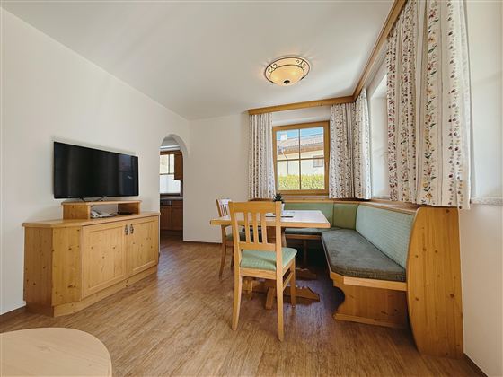 A bright and cheerful living room with a dining area and a television. Large windows allow plenty of daylight.