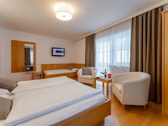 A modern hotel room with two beds and a cozy armchair. Large windows let in a lot of light and create a friendly atmosphere.
