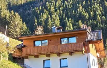 Ein modernes Haus aus Holz mit einem Balkon. Im Hintergrund erheben sich grüne Berge und Nadelbäume.