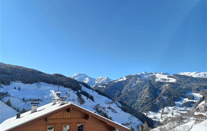 A picturesque mountain landscape with snow-covered hills and a clear blue sky. In the foreground stands a wooden house.