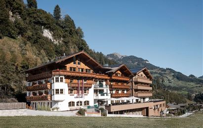 Ein gemütliches Hotel im Alpenstil, umgeben von grünen Wiesen und Bergen. Der Himmel ist klar und strahlend blau.
