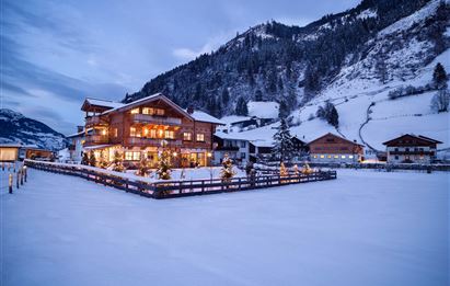 Eine gemütliche Berghütte im Schnee, umgeben von winterlicher Landschaft. Die Szene ist beleuchtet und zeigt einen klaren, bewölkten Himmel.