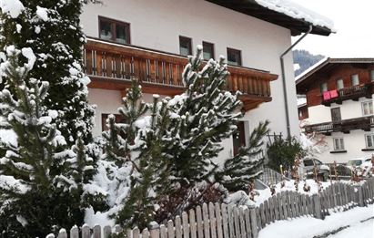 A winter scene with a house covered in snow. Green coniferous trees and a white fence complete the idyllic picture.